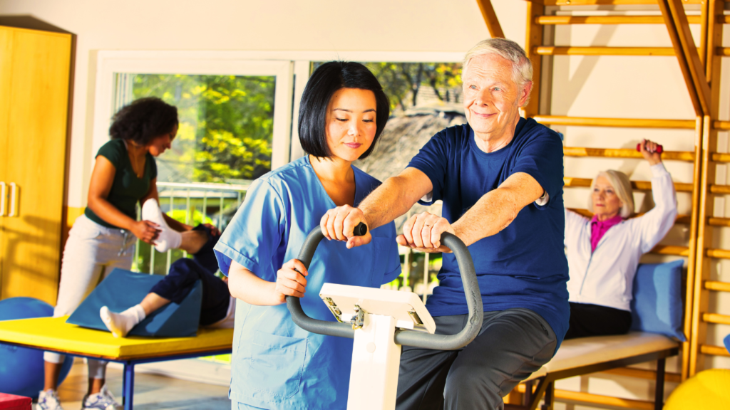 A Senior Man Exercising On A Static Bike With The Help Of A Rehabilitator 